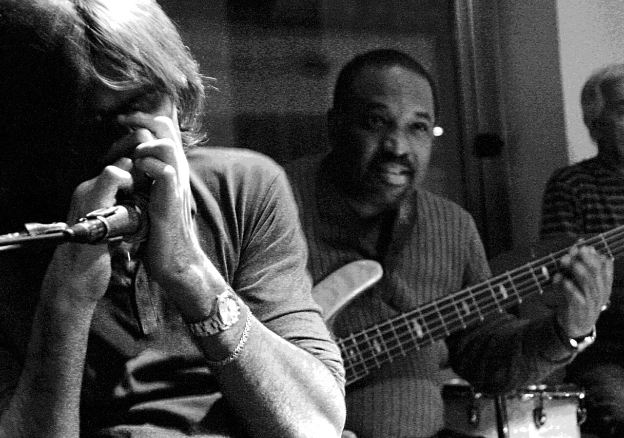 Músicos actuando en un club de jazz, uno toca armónica y otro guitarra.