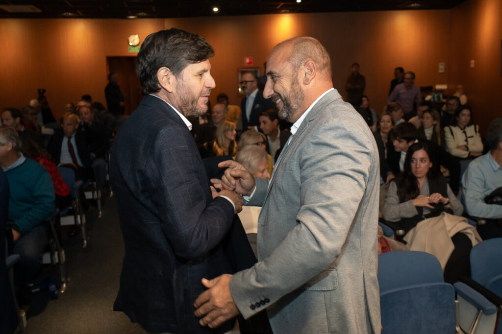 Dos hombres conversando en un evento con p&uacute;blico al fondo