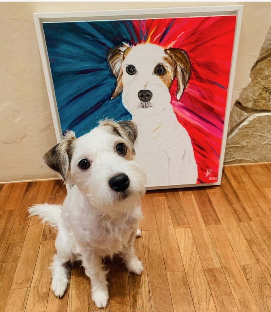 Perro blanco sentado frente a una pintura colorida de su retrato.