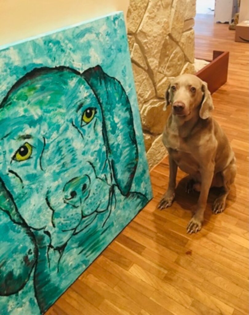 Perro sentado junto a una pintura de un perro azul