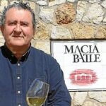 Ramon Servalls sosteniendo una copa de vino frente a un cartel de Macia Batle.