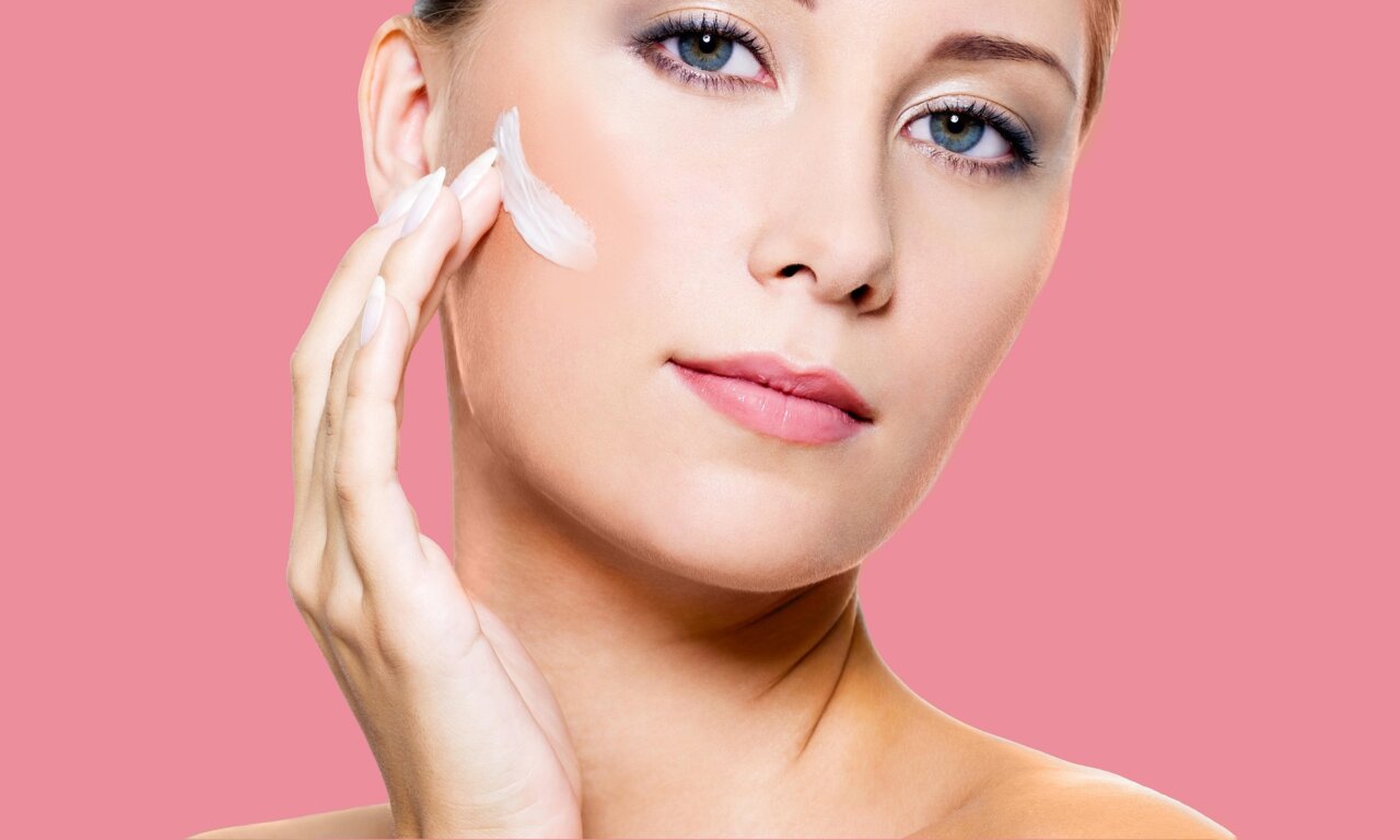 Mujer aplicando crema en su rostro con fondo rosa