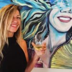 Maite Granados sosteniendo una copa de vino junto a su obra de arte
