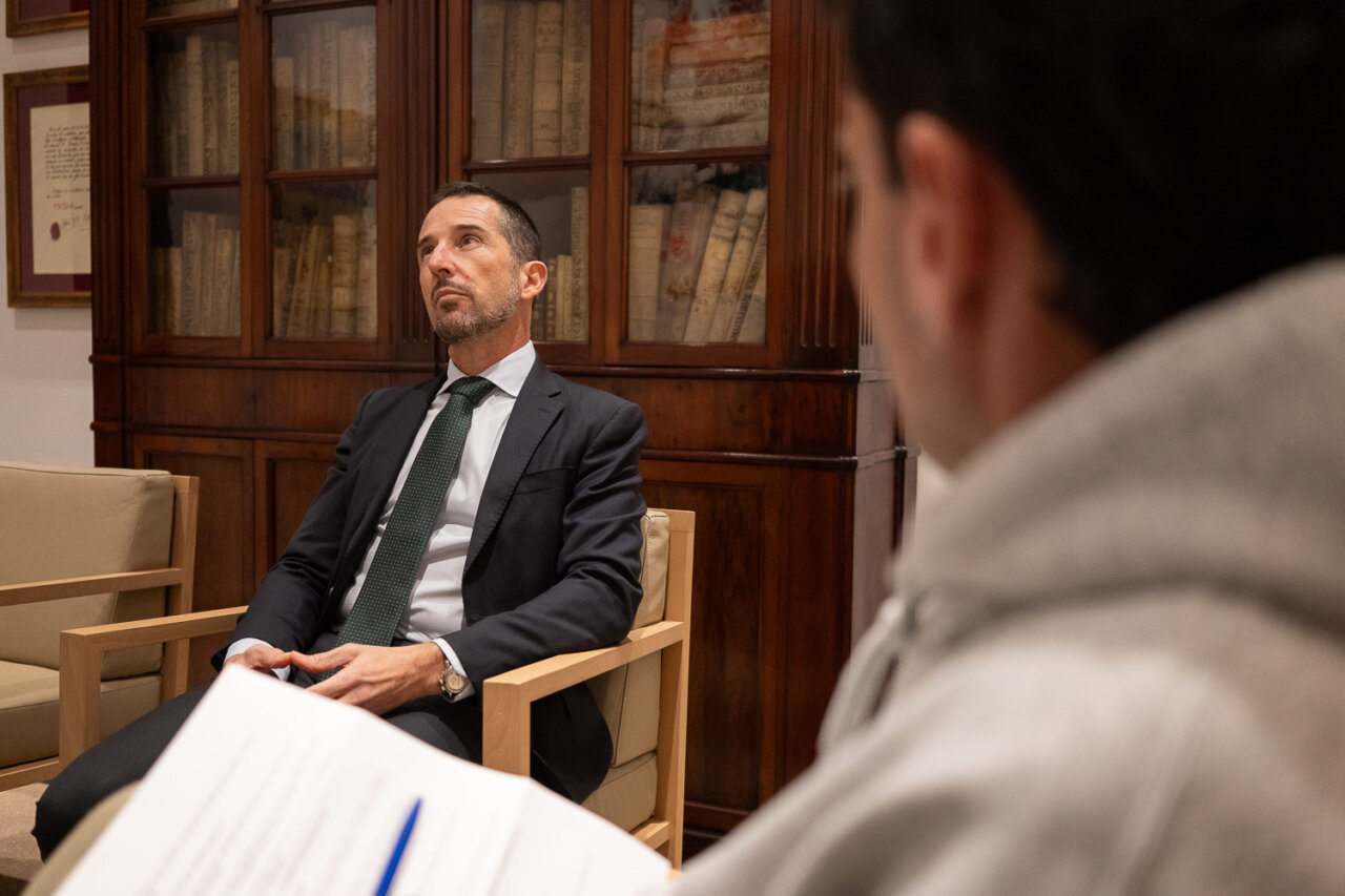 Hombre en traje sentado en una sala con estanterías de libros antiguos.
