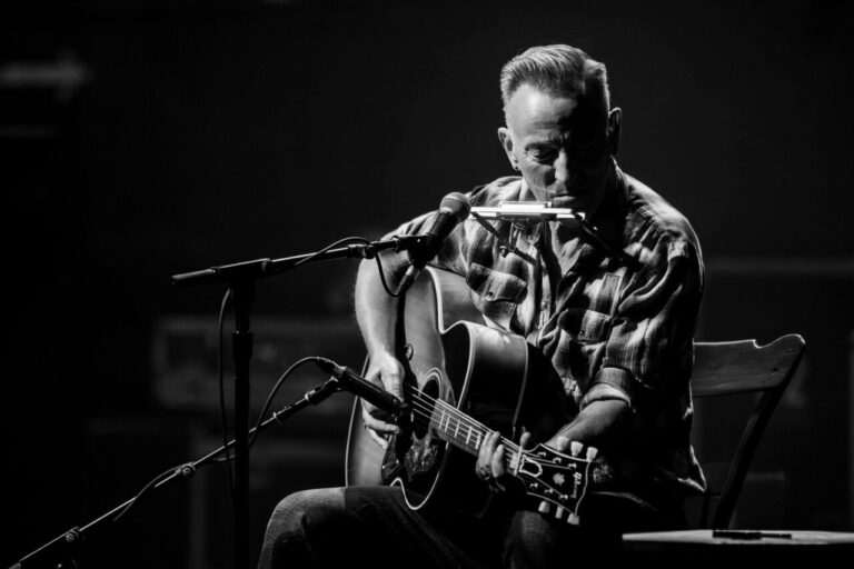 Bruce Springsteen tocando la guitarra y armónica en un escenario oscuro