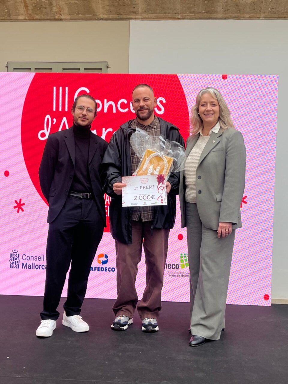 Tres personas posando en un evento de premiación con un cartel de primer premio.