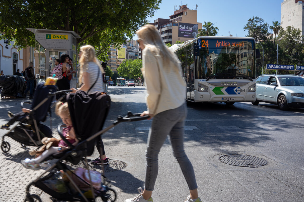 Personas cruzando la calle con carritos de bebé y un autobús en la parada.