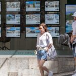 Mujeres caminando frente a una inmobiliaria con anuncios de vivienda