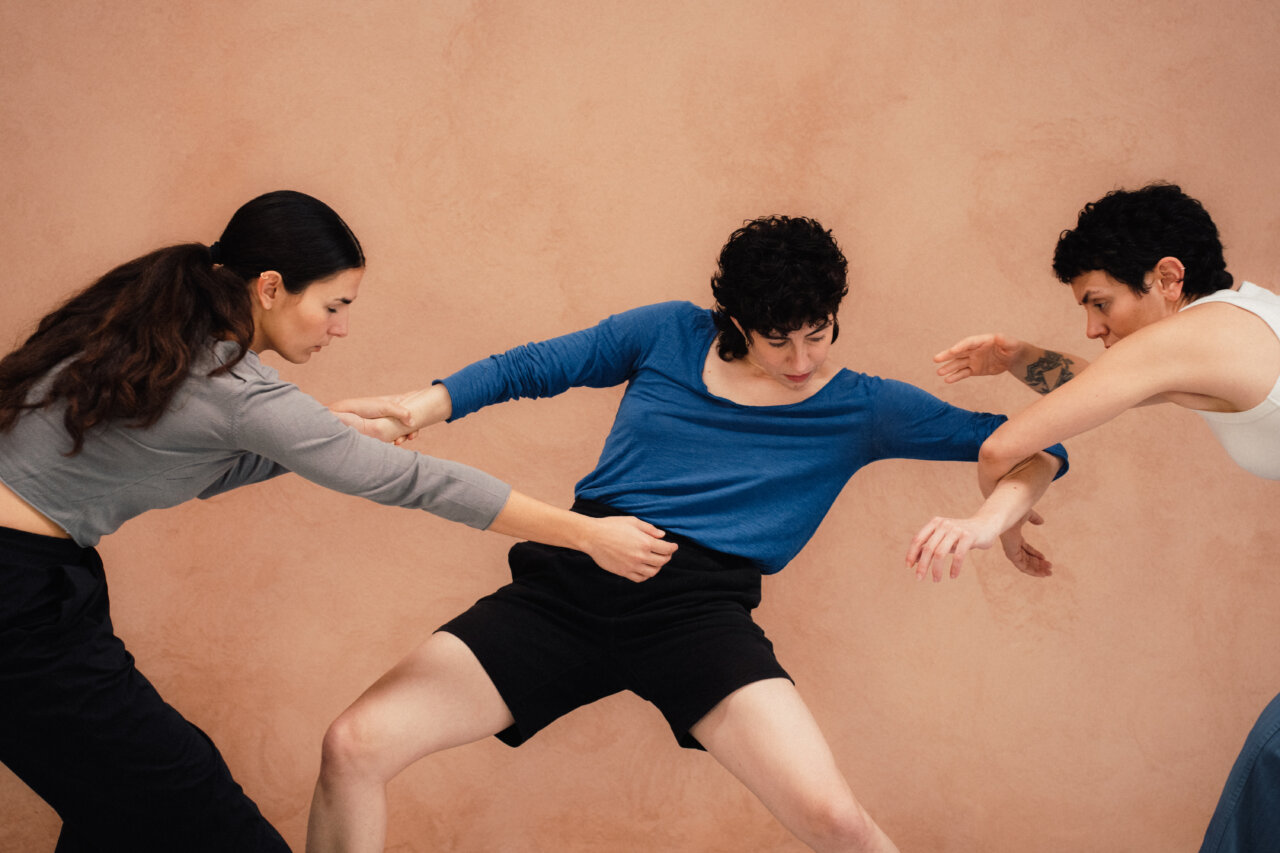 Tres bailarinas interactuando en una coreografía contemporánea en un fondo beige.