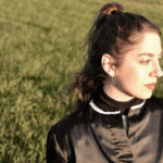 Maria Jaume en un campo verde, luciendo un atuendo elegante y reflexivo.