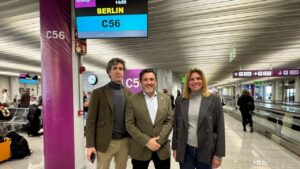 Tres personas en un aeropuerto con un cartel de vuelo a Berl&iacute;n