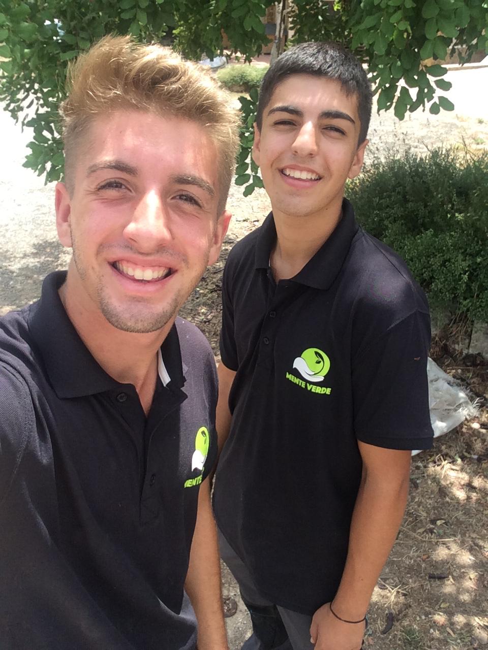Dos j&oacute;venes sonrientes en camisetas de jardiner&iacute;a bajo un &aacute;rbol