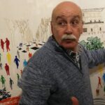 Hombre explicando un mural colorido con figuras humanas.