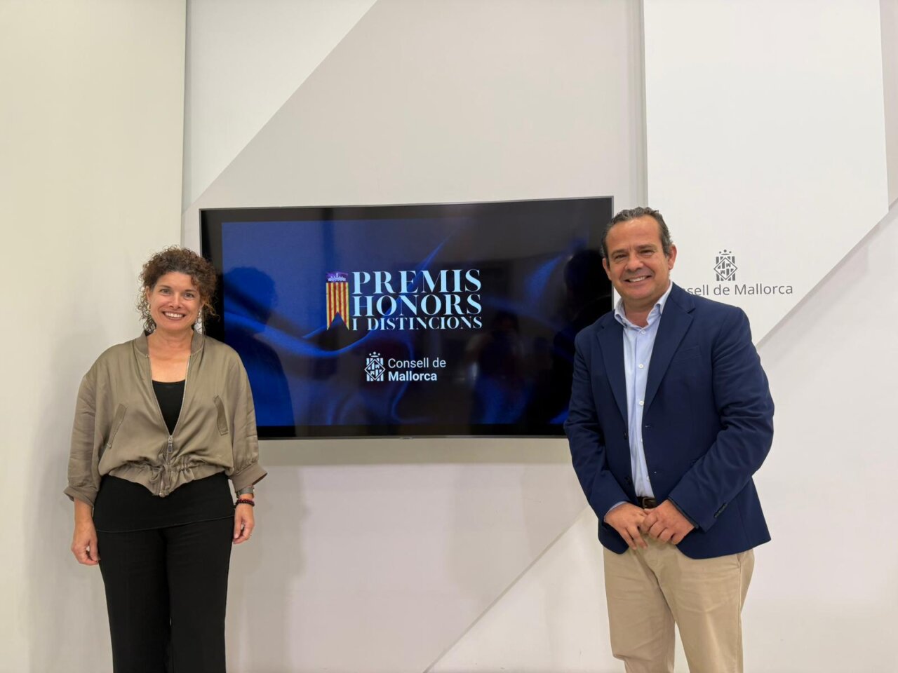 Dos personas posando frente a una pantalla sobre los Premis Honors i Distincions.