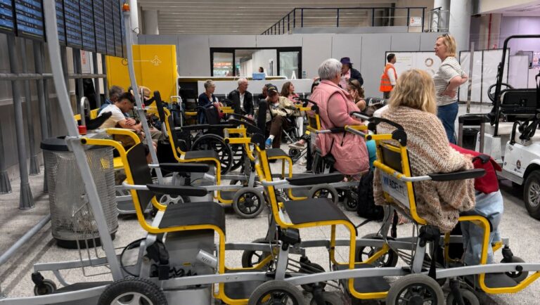 Varias sillas de ruedas en un aeropuerto con pasajeros esperando.