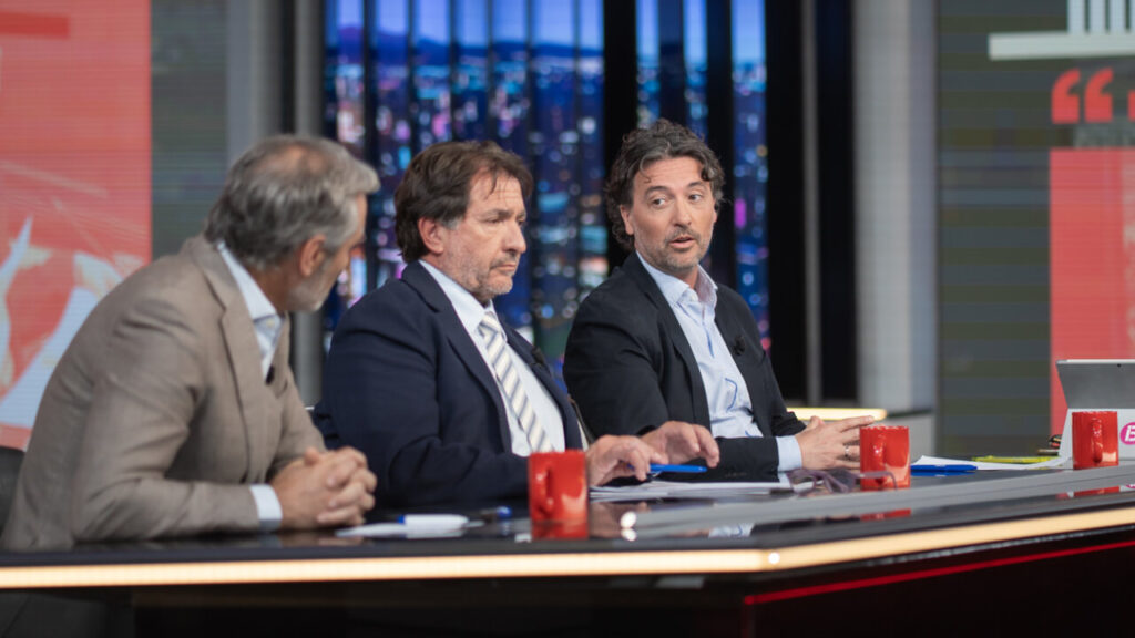 Tres presentadores discutiendo en un programa de televisión en un set moderno.