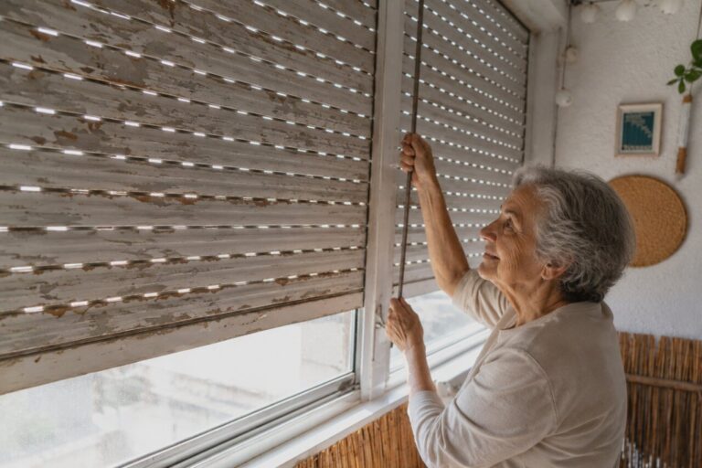 Mujer mayor ajustando una persiana en su hogar.