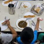 Ni&ntilde;os comiendo en un comedor escolar con platos vac&iacute;os y utensilios.
