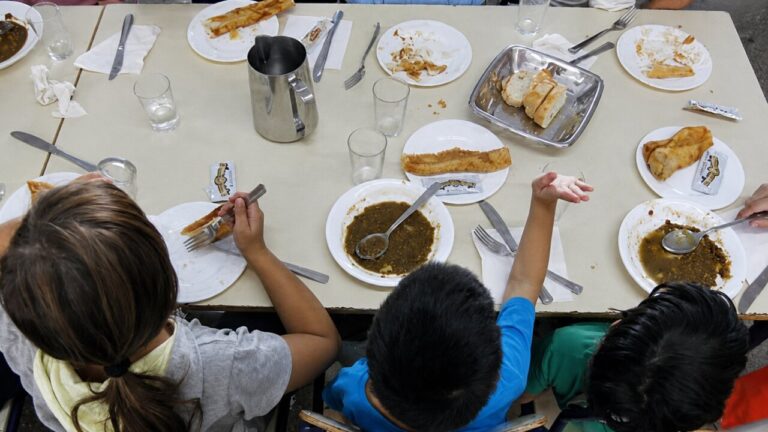 Ni&ntilde;os comiendo en un comedor escolar con platos vac&iacute;os y utensilios.