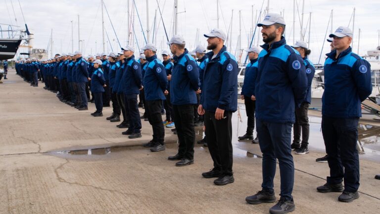 Grupo de agentes de Frontex alineados en un puerto con barcos al fondo