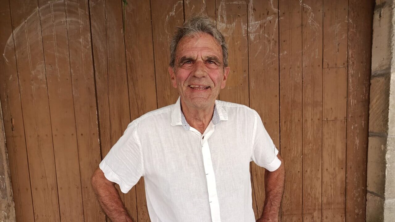 Hombre mayor con camisa blanca sonriendo frente a una puerta de madera