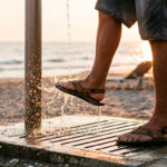 Persona lav&aacute;ndose los pies en una ducha de playa al atardecer