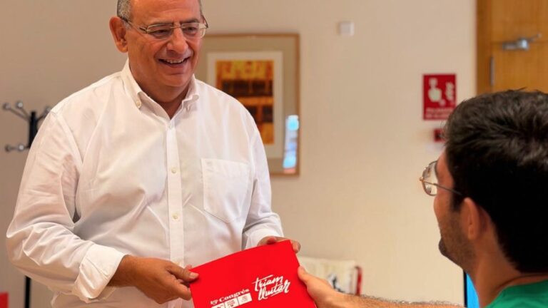 Alfonso Rodr&iacute;guez sonr&iacute;e mientras sostiene un documento rojo en una reuni&oacute;n.