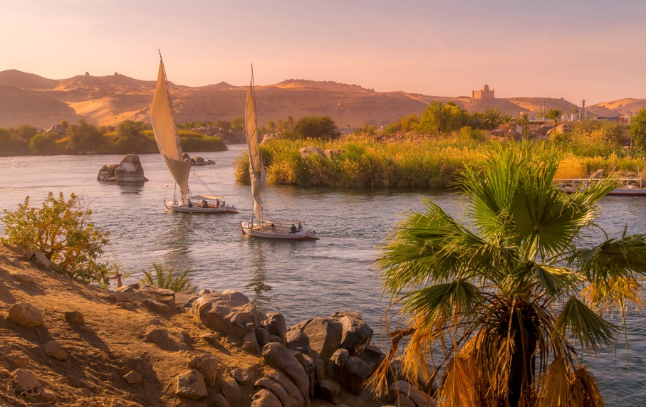 Vista de barcos de vela en un r&iacute;o rodeado de palmeras y monta&ntilde;as